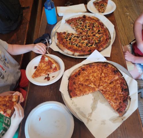 A table with three pizzas, one partially sliced, and two plates with pizza slices. Hands reach for the food.