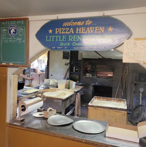 A cozy pizza shop interior with a welcoming sign, brick oven, and kitchen tools on a wooden counter.