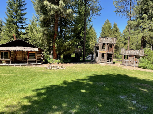 Two rustic wooden buildings and a sign in a grassy area surrounded by tall trees under a clear blue sky.