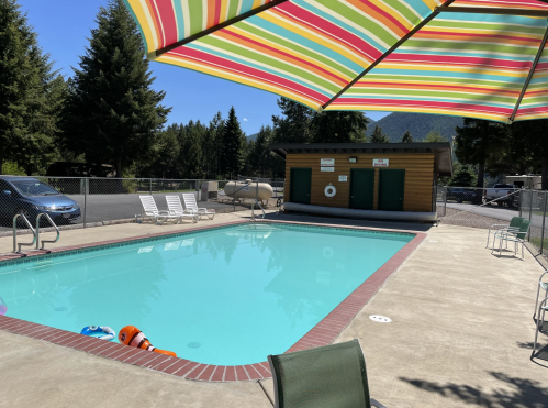 A sunny pool area with colorful umbrellas, lounge chairs, and a wooden building in the background surrounded by trees.