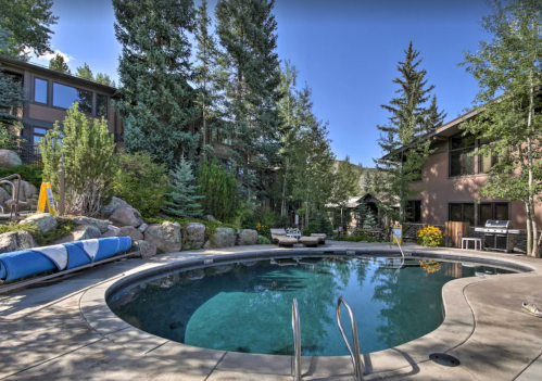 A serene outdoor pool surrounded by trees and rocks, with lounge chairs and a grill nearby.