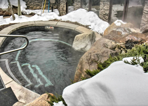 A steaming hot tub surrounded by snow-covered rocks and greenery, with a stone wall in the background.