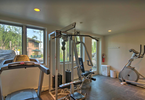 A bright gym with exercise equipment, including a treadmill and multi-gym, surrounded by large windows.