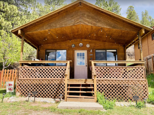 A wooden cabin with a porch, surrounded by greenery and decorative elements, under a clear blue sky.