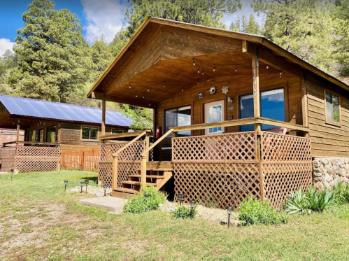 A cozy wooden cabin with a porch, surrounded by greenery and trees, featuring decorative lights and a nearby cabin.