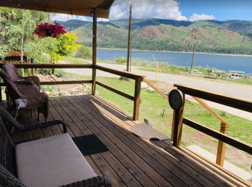 A wooden porch with seating overlooks a lake and mountains under a blue sky with fluffy clouds.