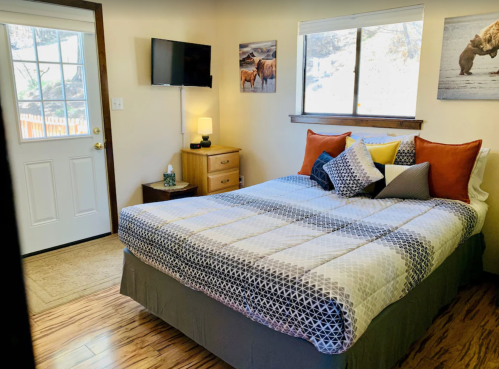 Cozy bedroom with a double bed, TV, wooden furniture, and artwork on the walls, featuring natural light from a window.