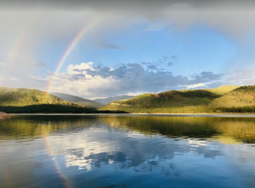 A serene lake reflects a vibrant rainbow and lush green mountains under a partly cloudy sky.