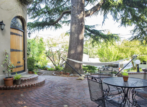 A cozy outdoor space with a brick patio, a large tree, a hammock, and a table set for dining.
