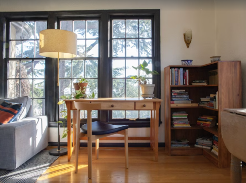 Cozy room with a wooden desk, chair, lamp, and bookshelves, featuring large windows and plants.