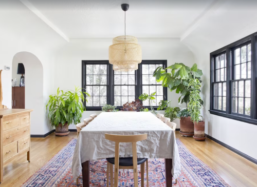 Bright dining room with a large table, woven chandelier, and lush plants by the windows, featuring a colorful area rug.