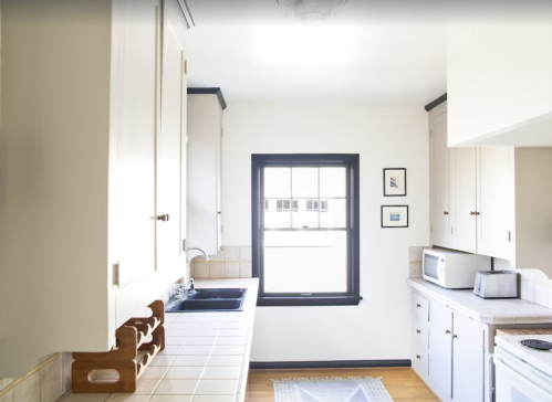Bright kitchen with white cabinets, a window, and a tiled countertop. Features a sink, microwave, and framed pictures.