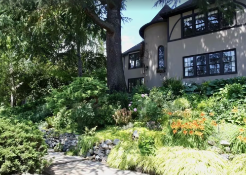 A charming house surrounded by lush greenery and colorful flowers, with a stone pathway leading to the entrance.