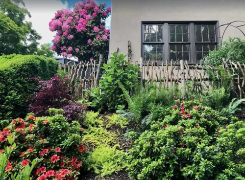 A lush garden with colorful flowers, greenery, and a wooden fence beside a house with large windows.