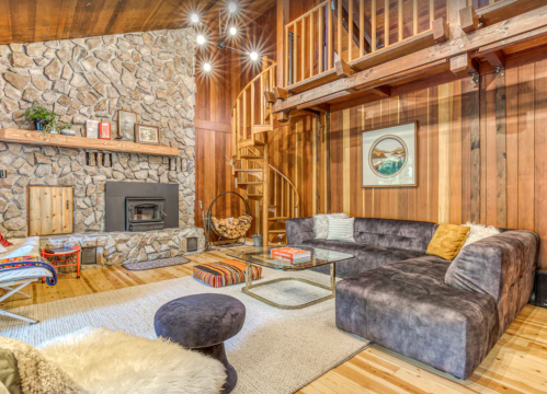 Cozy living room with stone fireplace, wooden walls, a sectional sofa, and a lofted area above. Warm and inviting atmosphere.