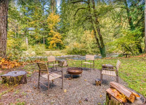 A serene outdoor campfire area with four chairs, a fire pit, and lush greenery by a river.