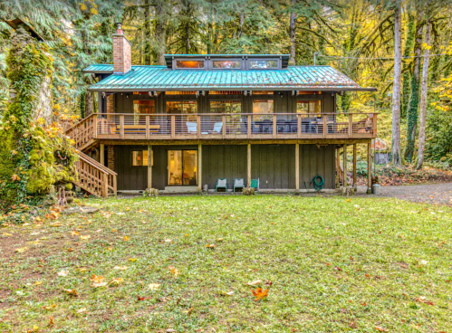 A modern two-story cabin surrounded by trees, featuring a green metal roof and a spacious deck with outdoor seating.