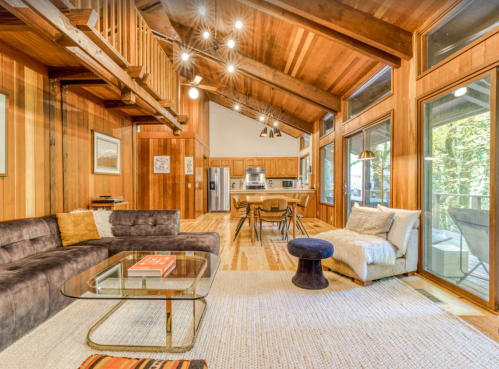 Cozy wooden interior of a modern living space with a sectional sofa, dining area, and large windows.