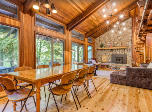 Cozy wooden dining area with a large table, stone fireplace, and large windows overlooking a forested view.