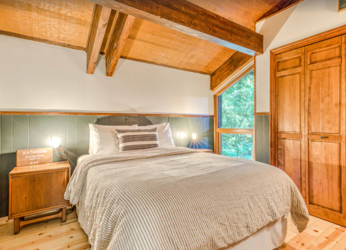 Cozy bedroom with wooden beams, a striped bedspread, and large windows overlooking greenery. Warm, inviting atmosphere.