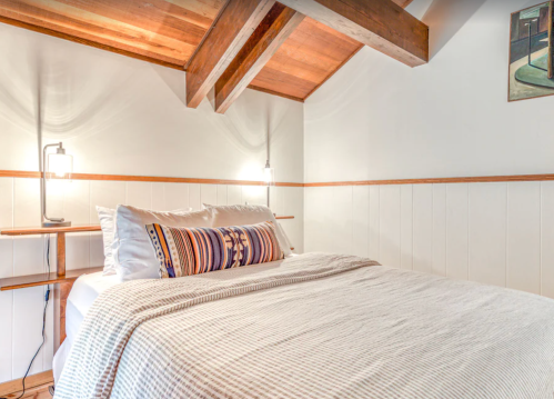 Cozy bedroom with a wooden ceiling, a neatly made bed, and soft lighting. Decor includes a patterned pillow and wall art.