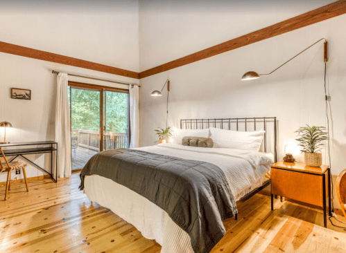 Cozy bedroom with a wooden floor, large window, and modern decor, featuring a bed, desk, and plants.