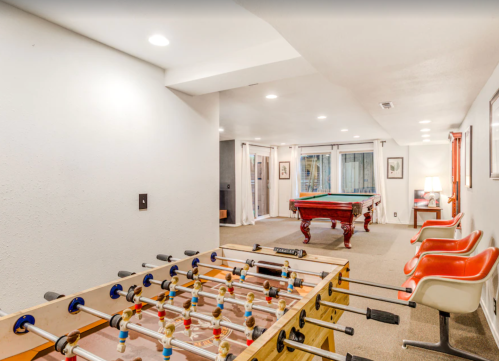 A game room featuring a foosball table, pool table, and seating area with large windows and neutral decor.