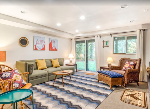 Cozy living room with a green sofa, colorful pillows, a woven chair, and large windows overlooking a garden.