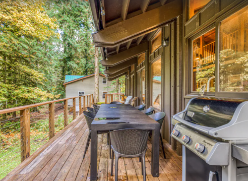 A wooden deck with a dining table and chairs, a grill, surrounded by trees and a cozy cabin in the background.