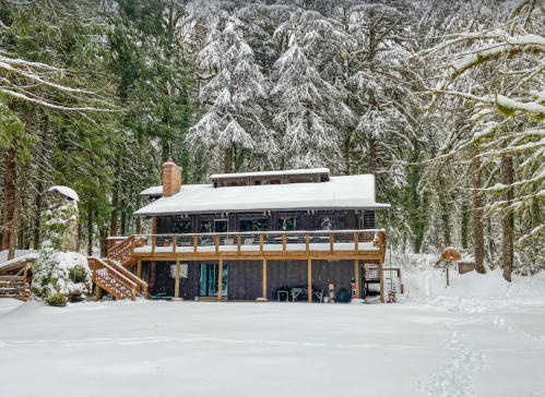 A cozy cabin surrounded by snow-covered trees, with a wooden deck and a snowy yard in a winter landscape.