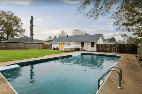 A backyard with a swimming pool, green lawn, and a house in the background under a cloudy sky.