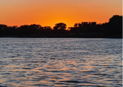 Sunset over a calm river, with silhouettes of trees against an orange and blue sky.