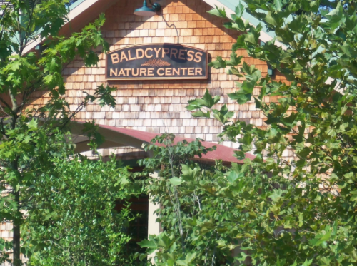 Sign for Baldcypress Nature Center on a wooden building, surrounded by greenery and trees.