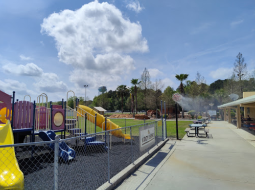 A colorful playground with slides and swings, surrounded by green grass and trees under a partly cloudy sky.