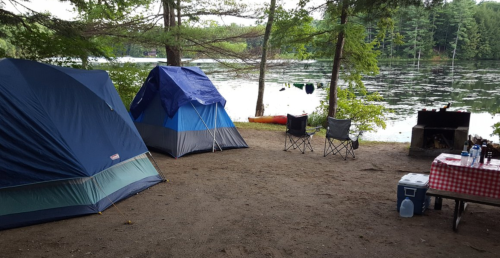 Two tents set up by a lake, with camping chairs and a picnic table nearby, surrounded by trees.