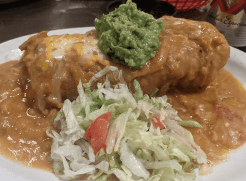 A large burrito topped with guacamole, served with a side of lettuce and tomato on a plate with sauce.