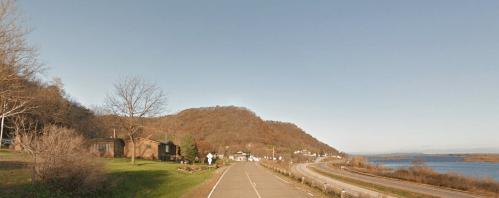 A scenic view of a road alongside a river, with a hillside and houses in the foreground under a clear blue sky.