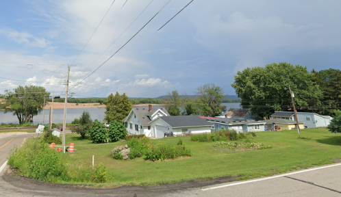A scenic view of a river with houses and greenery along the shore, under a cloudy sky. Power lines are visible.