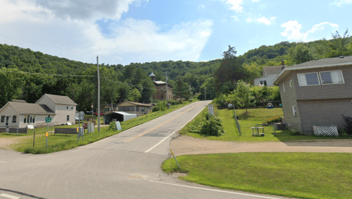 A quiet rural road lined with houses, leading up a hill with trees in the background under a partly cloudy sky.