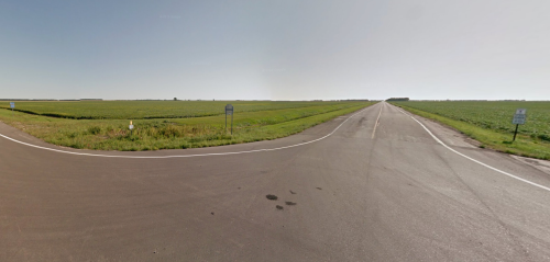 A rural intersection with a paved road splitting into two directions, surrounded by green fields under a clear sky.