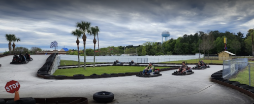 Go-kart track with racers navigating curves, palm trees in the background, and a water tower in the distance.