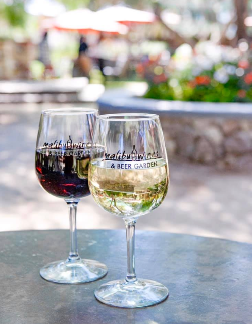Two wine glasses, one red and one white, sit on a table in a garden setting with colorful flowers in the background.