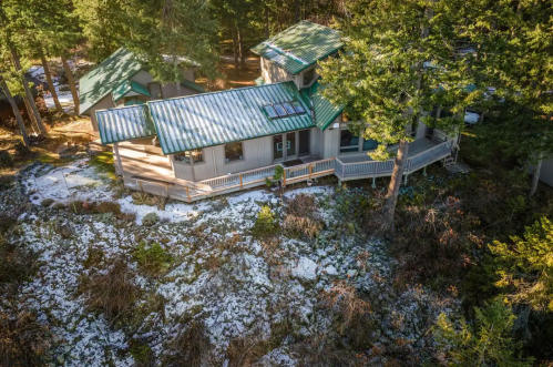 Aerial view of a modern house with a green metal roof, surrounded by trees and a snowy landscape.