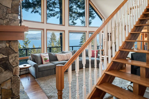 Cozy living room with large windows, a gray sofa, and a wooden staircase, offering a view of trees and a lake.
