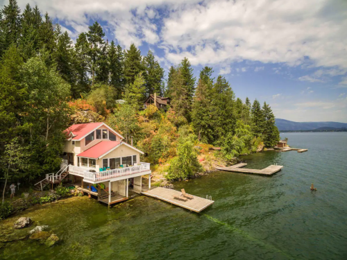 A lakeside house with a red roof, surrounded by trees, featuring docks extending into the water under a blue sky.
