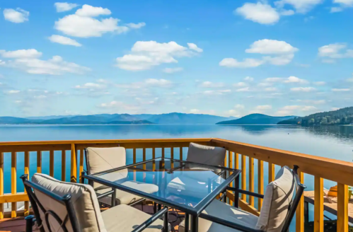 A scenic view of a calm lake from a wooden deck with a glass table and chairs, under a blue sky with fluffy clouds.