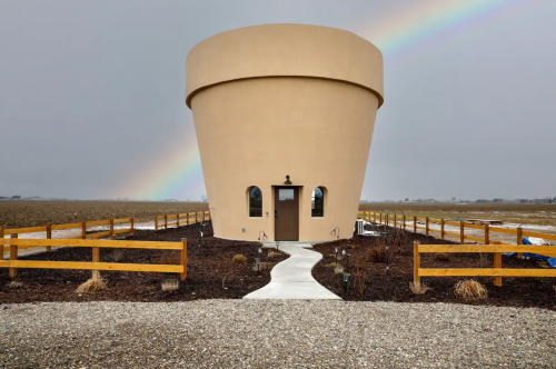 A unique, round building resembling a flower pot, with a rainbow in the background and a path leading to the entrance.
