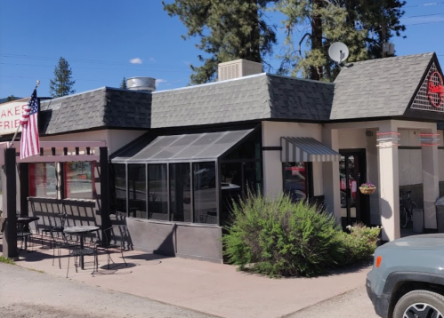 A small restaurant with a gray roof, outdoor seating, and an American flag, surrounded by trees and blue sky.