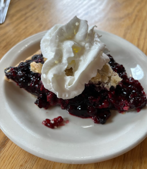A slice of berry pie topped with whipped cream on a white plate, resting on a wooden table.