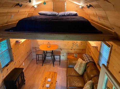 Cozy wooden interior of a tiny home with a lofted bed, dining table, and seating area. Warm, inviting atmosphere.